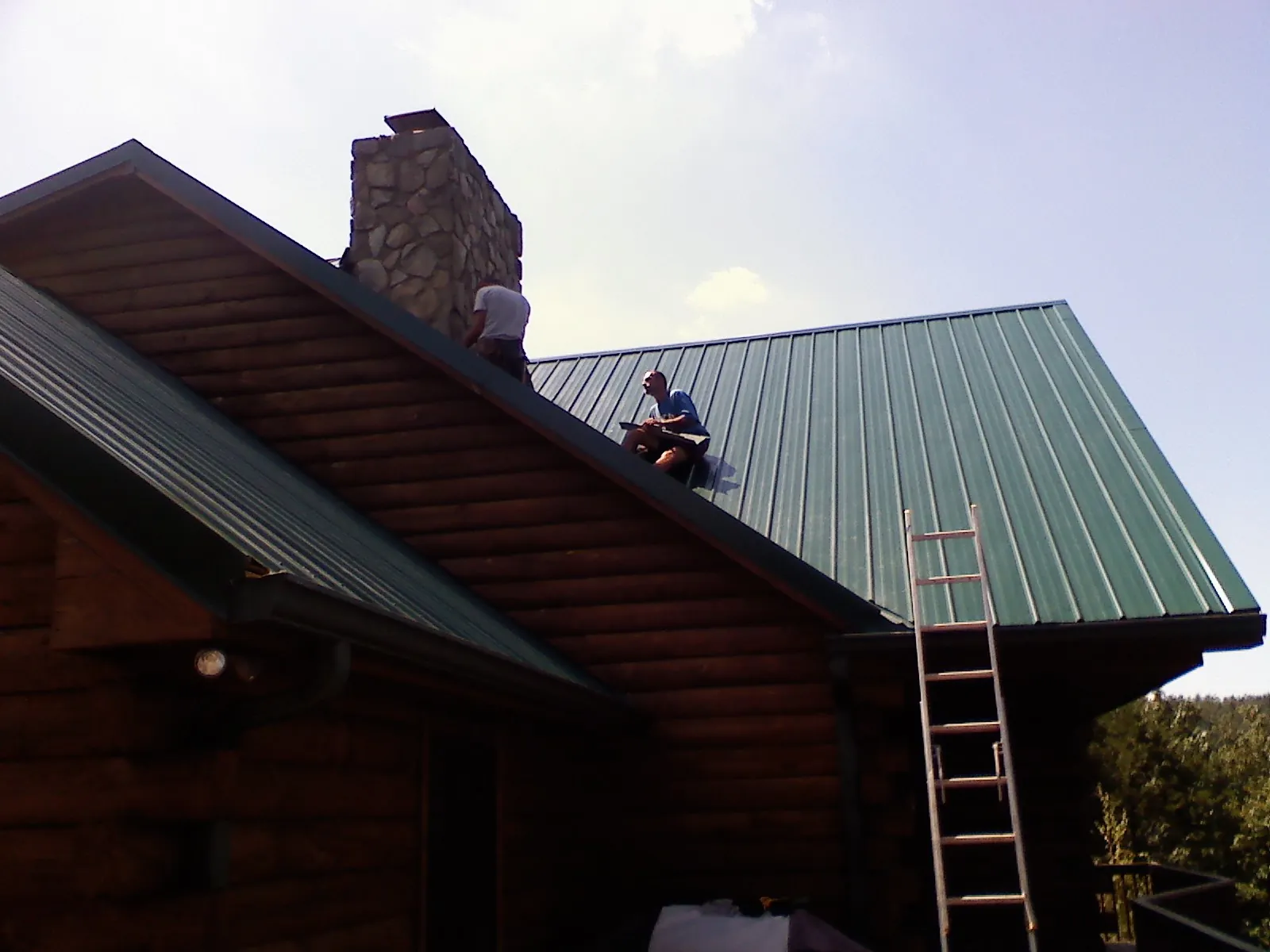 Roofers installing green metal panels beside a stone chimney on a log home in Agate
